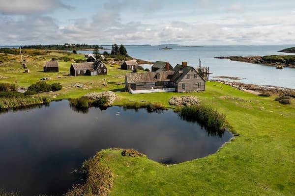 An elevated landscape photograph capturing a cluster of weathered wooden houses on a grassy, rocky island. A dark, calm pond sits in the foreground, reflecting a soft blue sky filled with white clouds. In the background, the rolling green land meets a calm ocean cove dotted with distant islands.
