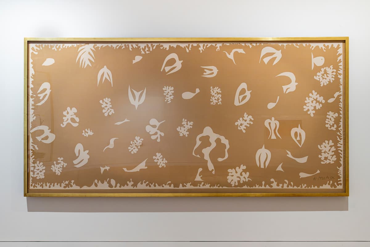 A large, horizontally oriented artwork by Henri Matisse installed on a white gallery wall. The piece features a muted tan or beige background filled with white silk-screened cutouts of stylized marine life, birds, coral, and organic shapes. The artwork is enclosed in a simple, thin gold frame.