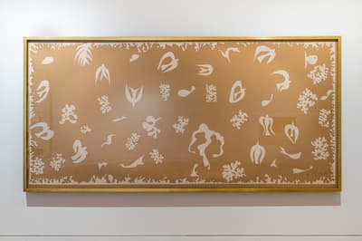 A large, horizontally oriented artwork by Henri Matisse installed on a white gallery wall. The piece features a muted tan or beige background filled with white silk-screened cutouts of stylized marine life, birds, coral, and organic shapes. The artwork is enclosed in a simple, thin gold frame.