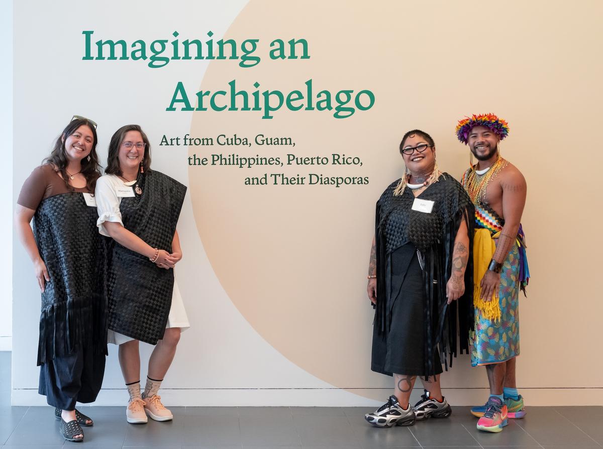Four artists smiling for a photo in front of a wall with the exhibition title, "Imagining an Archipelago: Art from Cuba, Guam, the Philippines, Puerto Rico, and Their Diasporas".