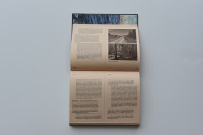 An open multi-page booklet or catalog rests flat against a light-grey background. The booklet is open to a double-page spread printed on tan-colored paper. The left page features printed text columns in English, while the right page shows two small black-and-white landscape photographs of a quiet country road alongside text in other languages.