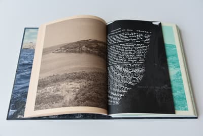 n open multi-page book or catalog rests flat against a light-grey background. The left page is printed on tan paper and features a sepia-toned landscape photograph of a grassy shoreline curving around a calm body of water. The right page is stark black, featuring stylized white typography overlaid with the dark silhouette of a palm tree. The outer edges of the open book reveal glimpses of its endpapers, displaying a blue sea landscape on the left and a vibrant teal-tinted sea texture on the right.