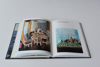 An open multi-page book or catalog rests flat against a light-grey background. The book is open to a double-page spread featuring two colorful paintings. The left page displays a figurative painting of a group of people gathered around a dining table inside a room with yellow curtains. The right page features a still life painting of a table set with dishes and a pitcher on a green-and-white checkered tablecloth against a textured blue wall, with small printed caption text above. The dark-blue sea texture of the book's outer cover is visible along the left edge.