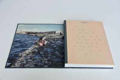 An open book or catalog is presented flat against a light-grey background, showing the inside front cover and first page. The left side (inside cover) features a full-bleed color photograph of a person crouching on a rugged, dark rocky shore next to the blue ocean, with stone fortifications in the background under a blue sky. The right side is a tan-colored page with a dark-grey spine edge. It features minimalist black lettering scattered across the surface, spelling out "archipelagos resemble constellations" in a sparse, constellation-like layout.