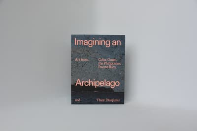 A printed booklet or catalog standing vertically against a solid, light-grey background. The cover features a graphic design with a textured, split-tone background of dark teal and deep ocean blue. Peach-colored typography overlays the surface, reading "Imagining an" at the top, "Art from Cuba, Guam, the Philippines, Puerto Rico," across the middle, "Archipelago" in bold, large font near the bottom, and "and Their Diasporas" along the lower edge.