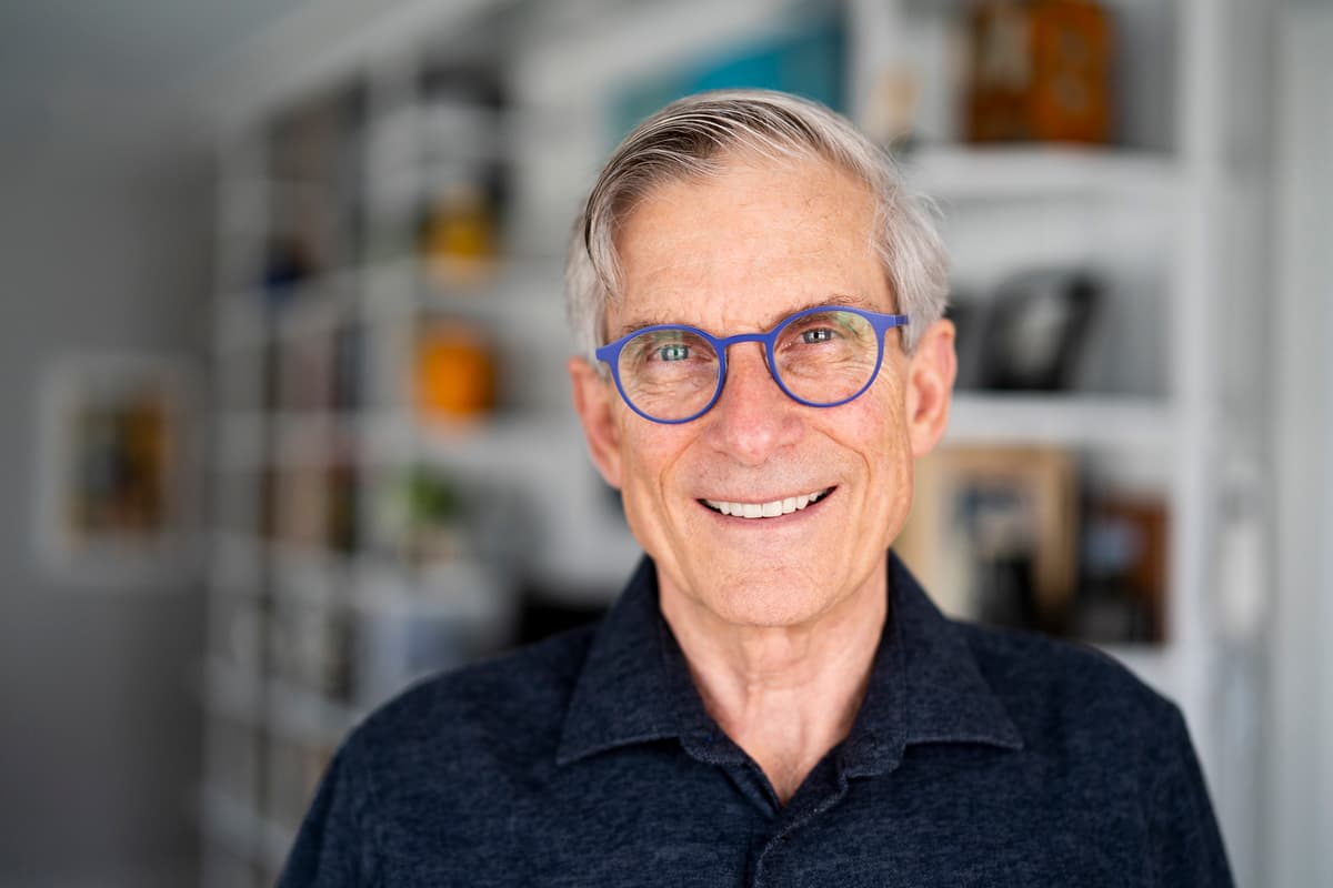 Headshot of a smiling older man with short graying hair and blue-rimmed glasses. He is wearing a dark, textured button-up shirt and standing in front of a softly blurred white bookshelf background.