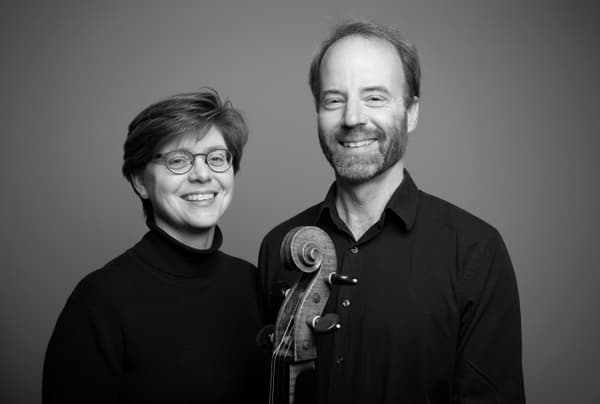 Black-and-white studio portrait of a smiling man and woman side by side. The woman, wearing glasses and a black turtleneck, stands on the left. The man, with a light beard and dark button-down shirt, holds the scroll and neck of a cello in front of them.