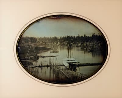 Oval-framed vintage daguerreotype or early photograph showing a river scene with floating bamboo bridges and small boats. Traditional thatched-roof dwellings line the distant shoreline along the calm water, framed within a decorative brass and cream mat.