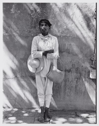 Black and white portrait of a barefoot man standing outdoors with arms crossed against a textured wall dappled with tree shadows. He wears light-colored, thin-striped long clothing, holding a wide-brimmed hat under one arm and a woven shoulder bag over the other.