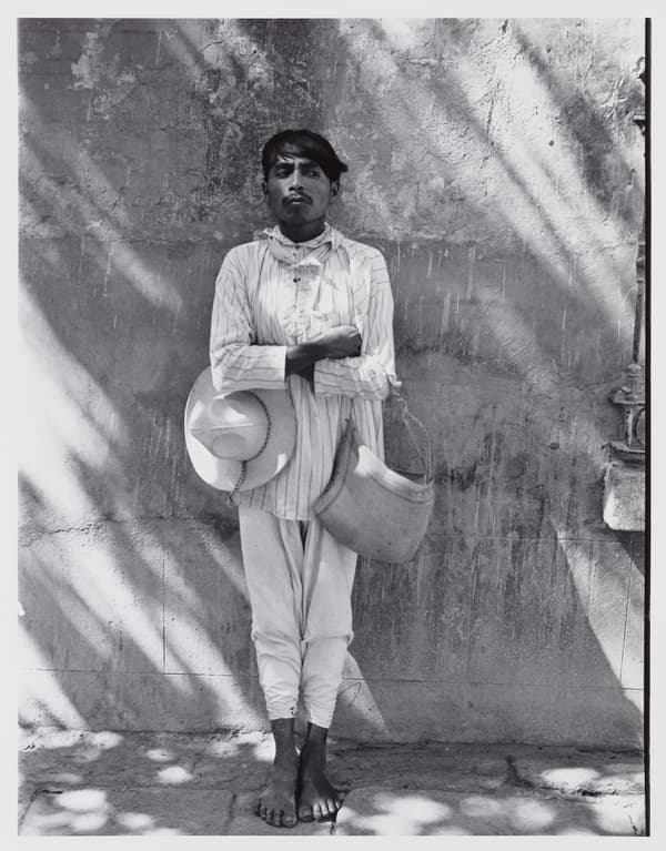Black and white portrait of a barefoot man standing outdoors with arms crossed against a textured wall dappled with tree shadows. He wears light-colored, thin-striped long clothing, holding a wide-brimmed hat under one arm and a woven shoulder bag over the other.