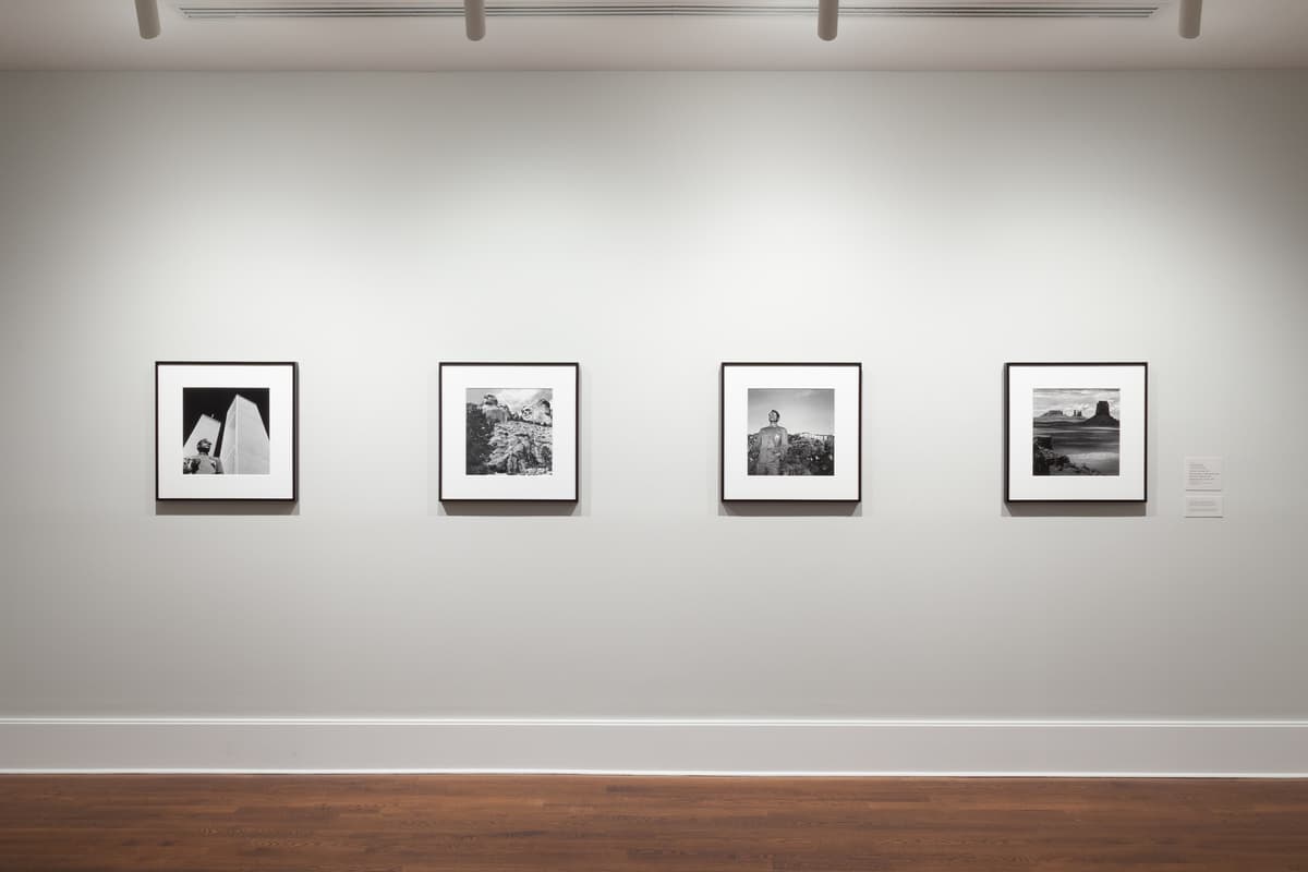 Gallery with four square black-and-white photographs mounted onto a grey wall in a room with wooden.