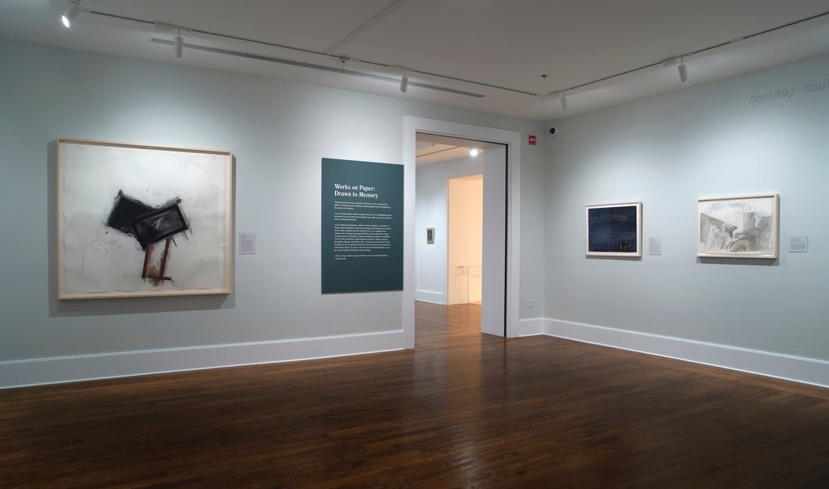 Corner gallery view featuring a dark teal exhibition text panel titled "Works on Paper: Drawn to Memory" situated next to an open doorway. To the left of the doorway hangs a large abstract drawing, while two smaller framed artworks hang on the adjacent wall to the right above dark hardwood floors.