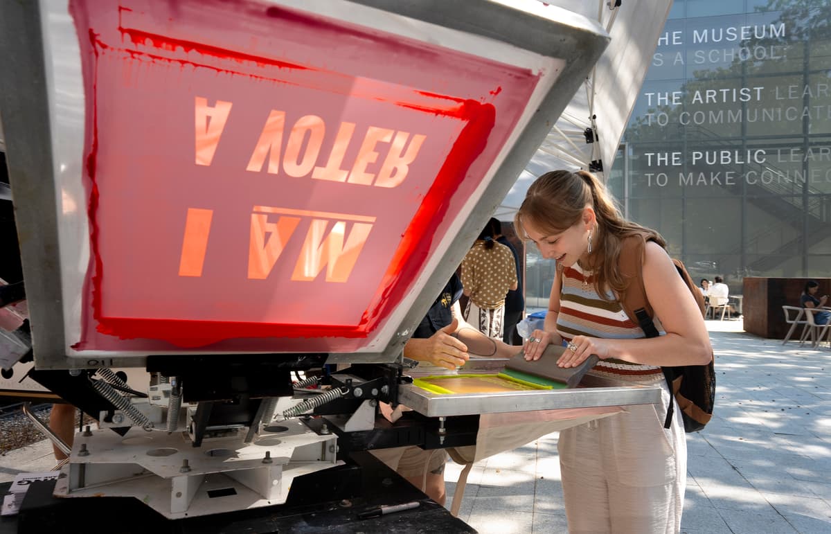 Person screenprinting in front of the museum by a larger screenprint screen reading, "I am a voter".