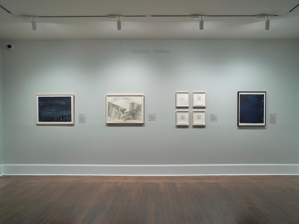Gallery installation view featuring four groupings of framed artworks mounted on a light gray wall beneath subtle text reading "Gourley Gallery." The wall displays two horizontal landscapes on the left, a two-by-two grid of small square prints in the center, and a dark vertical painting on the right above dark hardwood floors.