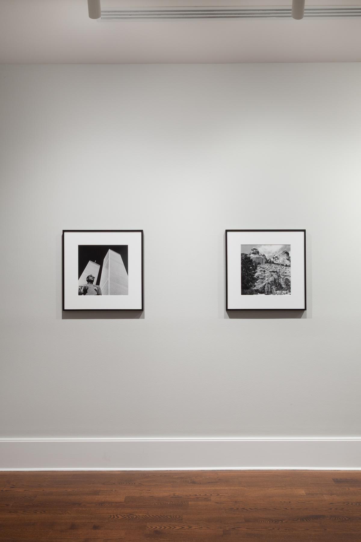Installation view of two framed black and white photographs hung in a gallery on grey walls with wooden floors. On the left, a figure is visible in front of the World Trade Center towers and on the right Mount Rushmore.