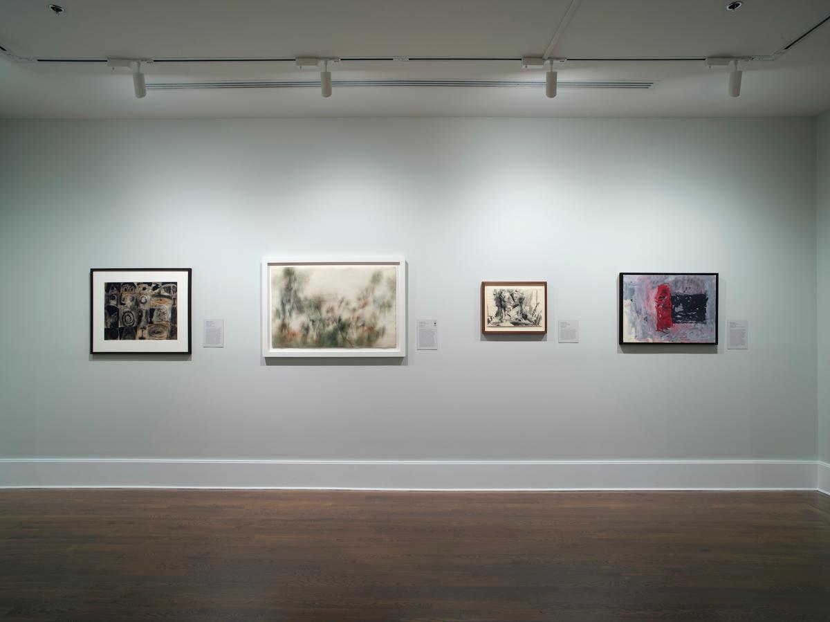 Installation view of four varied framed paintings hung in a horizontal line along a smooth light gray gallery wall above dark wood floors and white baseboards, with track lighting mounted on the ceiling above.