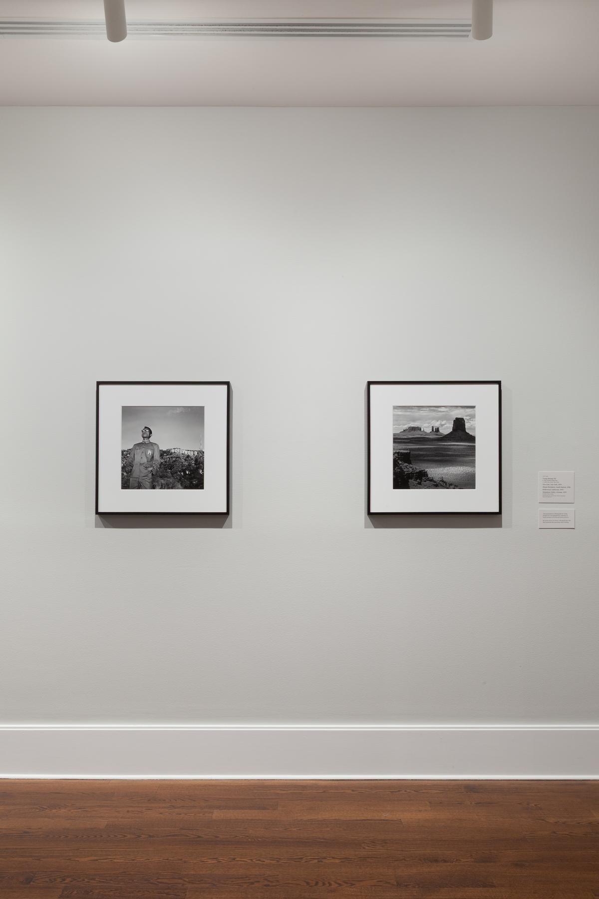 Installation view of two framed black and white photographs hung in a gallery on grey walls with wooden floors. Installation view of two framed black and white photographs hung in a gallery on grey walls with wooden floors. On the left, a figure is visible in front of the Hollywood sign and on the right a desert landscape.