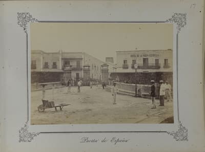 Historic sepia-toned photograph mounted in an ornate paper border depicting a city entrance labeled "Puerta de España" in San Juan, Puerto Rico. People in 19th-century attire gather in an open paved plaza area near stone gateposts, with a wooden handcart in the foreground and a multi-story building marked "HOTEL DE LA NUEVA ESTRELLA" visible in the background.