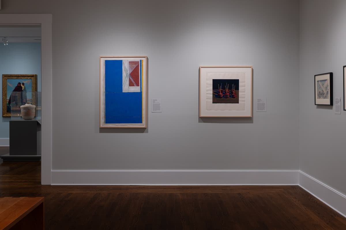Art gallery interior view featuring framed artworks displayed on light gray walls above dark hardwood floors. On the main wall, a large vertical abstract piece with a bold blue color field hangs next to a framed print depicting red candied apples on sticks. Through a doorway to the left, another gallery room displays a painting and a ceramic vase on a pedestal.