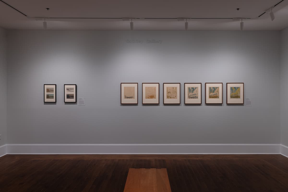 Two print series, one a pair and the other a series of five works, framed on a gray wall in a gallery with grey walls, wooden floors, and a bench.