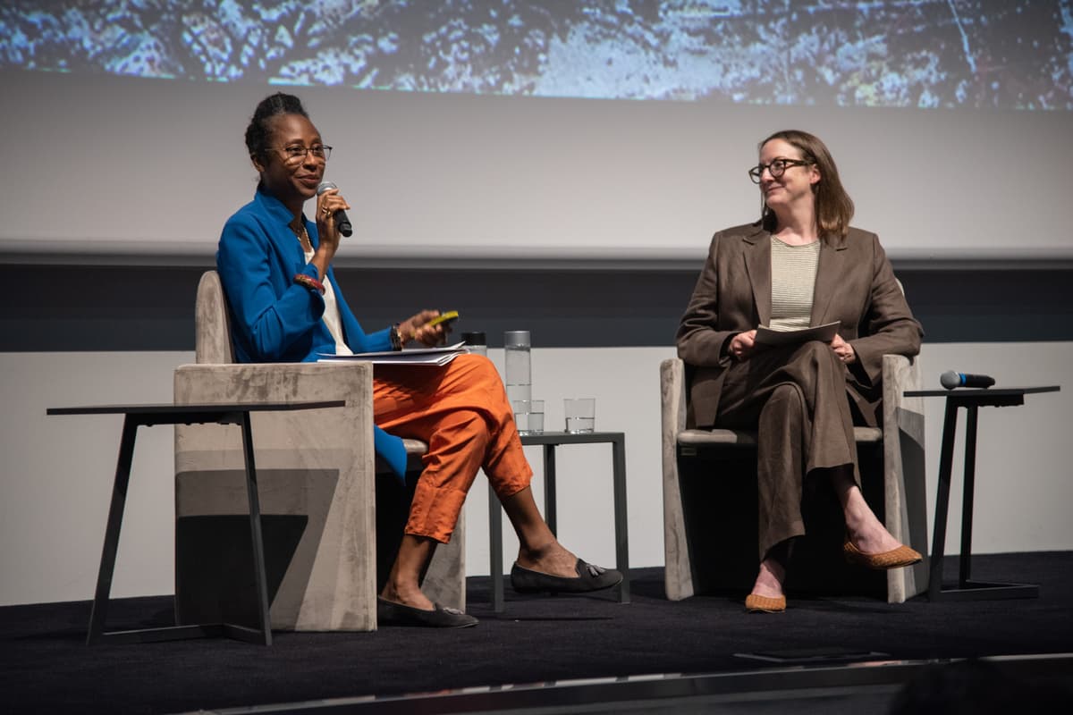 Naomi Beckwith and Lucy Bradnock seated in armchairs on stage during a presentation. Beckwith, wearing a vibrant blue blazer and bright orange trousers, speaks into a microphone while holding a clicker. Bradnock, in a brown suit and glasses, listens smilingly with notes in her lap.