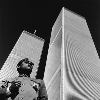 Black and white portrait of artist Tseng Kwong Chi standing in front of the World Trade Center twin towers in New York City.