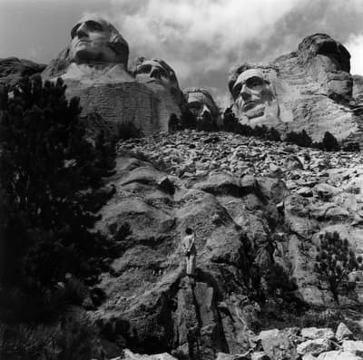 Black-and-white photograph of Mount Rushmore shot from beneath. In the center foreground a figure with their back to the camera looks up at the sculpture.