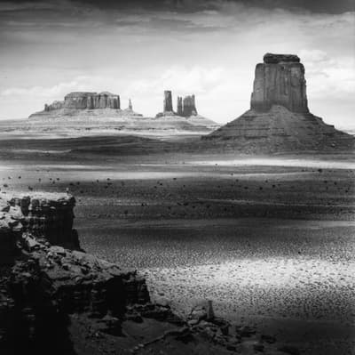 Black-and-white photograph of the Monument Valley desert landscape. Center left a figure is faintly visible on a cliff.