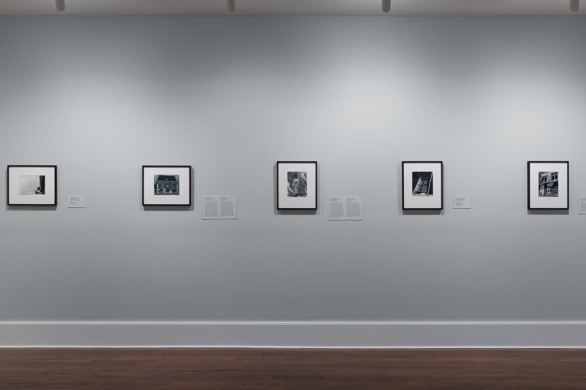 Gallery wall view featuring five black-and-white photographs by Manuel Álvarez Bravo, uniformly framed and spaced in a horizontal row across a smooth light gray wall above white baseboards and dark hardwood flooring. Each artwork is accompanied by a white wall label to its right.