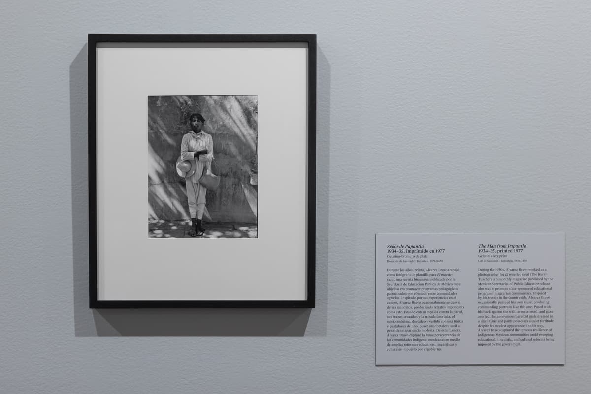 Framed black-and-white portrait by Manuel Álvarez Bravo hanging on a gray gallery wall next to an informational didactic label. The photograph captures a barefoot man standing with arms crossed against a wall with dappled shadows, wearing light attire and holding a wide-brimmed hat.
