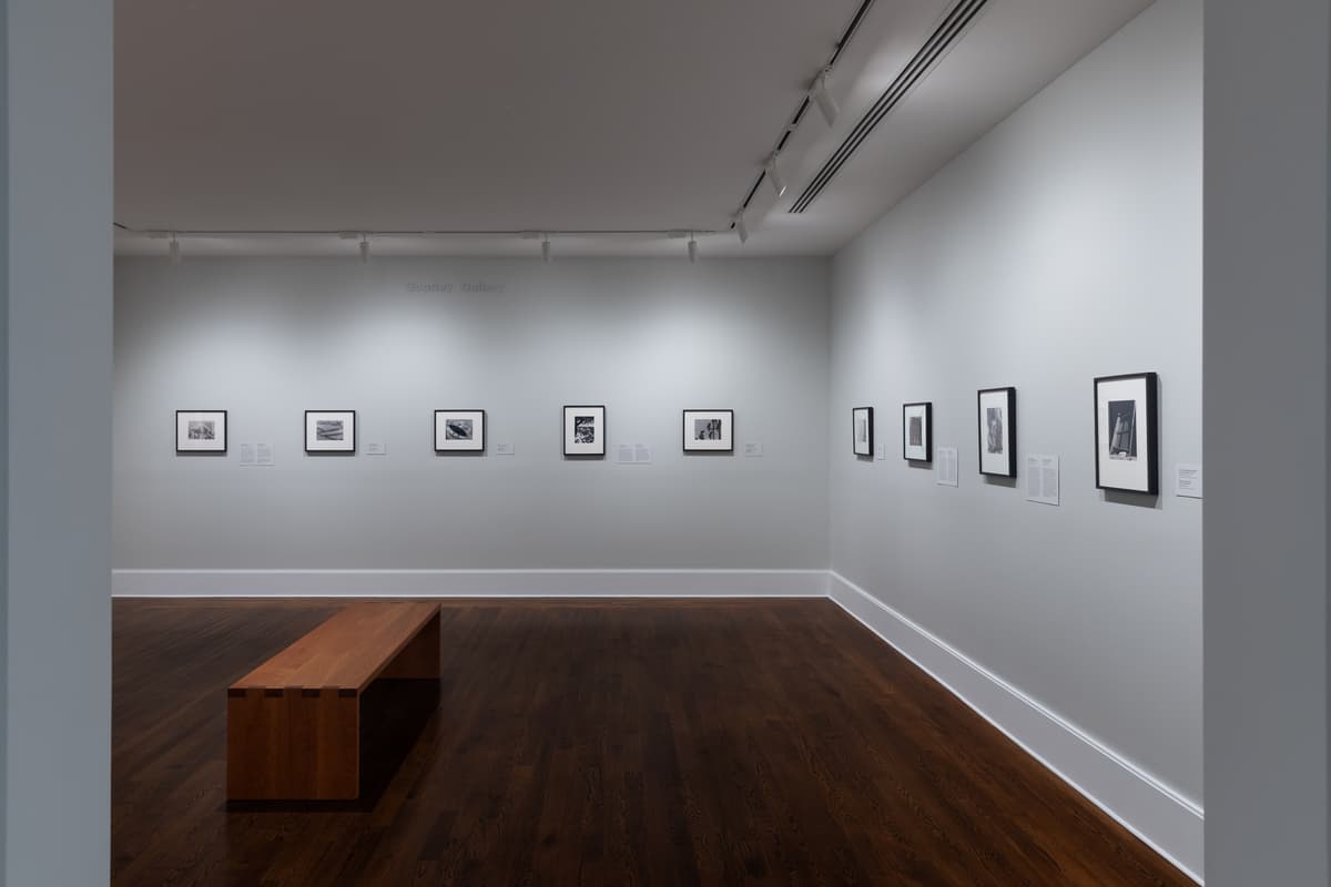 Art gallery installation view featuring small, framed black-and-white photographs hung in a precise horizontal row along light gray walls. A wooden wooden bench sits on the dark hardwood floor in the foreground, facing the far wall under track lighting.