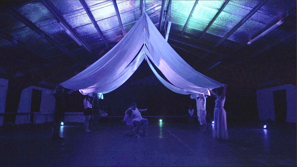 Gi Matan Guma' performers holding up white cloth to form a roof over a performer seated in a chair.