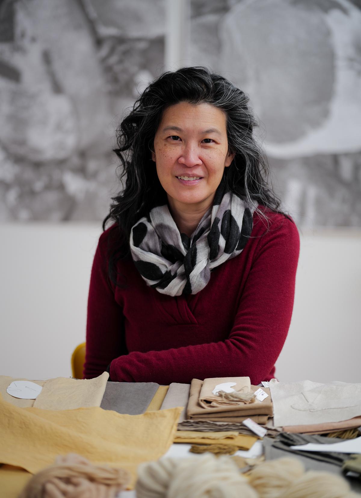 Portrait of artist Elaine K. Ng smiling at the camera, seated behind a table covered in neutral-toned textiles and yarn swatches. She wears a deep red sweater and a patterned scarf, with black-and-white artwork blurred in the background.