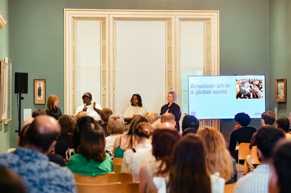 A panel of four speakers seated on stage during a public talk titled "American art in a global world." The speakers face a seated audience in a grand room with sage-green walls, white-trimmed arched windows, and a large display screen showing the presentation title on the right.