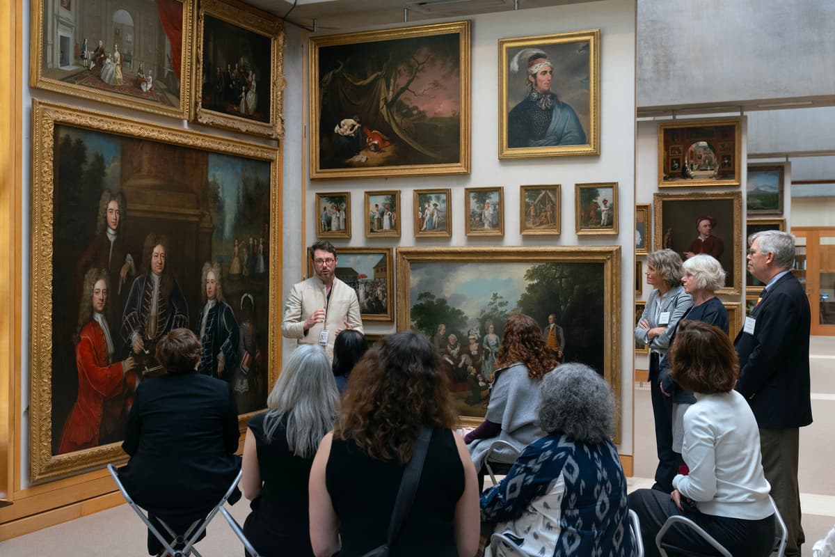 A museum guide stands in a gallery gesturing toward a wall filled with salon-hung oil paintings in ornate gold frames, leading a tour group seated in folding chairs and standing around him.