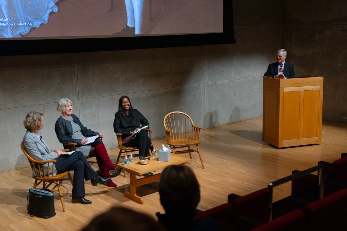 Three women sit in wooden chairs on a stage during a panel discussion, while a man speaks from a podium on the right. A large screen behind them displays artwork, and an empty wooden armchair sits next to a coffee table on stage.