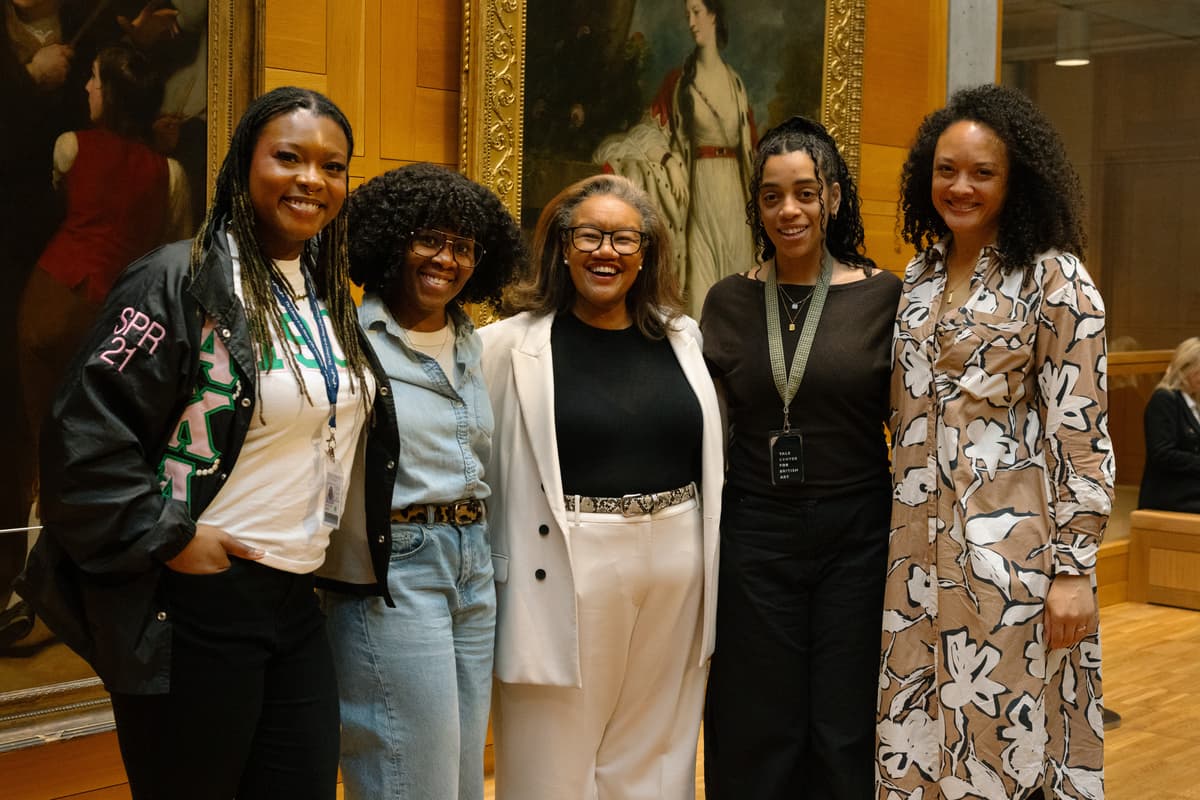 Five smiling women pose together in front of a framed classical portrait hanging on a wooden wall inside an art museum.