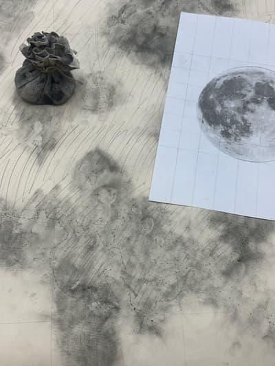Image of a moon on paper and material used for spolvero on a surface covered in dark pigment.