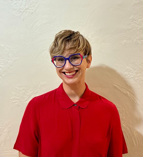 Studio headshot of a smiling person with short, textured wavy blonde-and-brown hair, vibrant blue-and-pink eyeglass frames, and a nose ring. They are wearing a collared, short-sleeved bright red shirt, posed against a textured, light beige wall.
