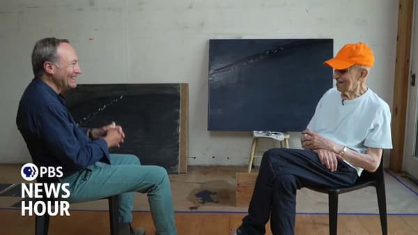 Two men sit facing each other in chairs inside a brightly lit art studio, conversing. The man on the left, Jeffrey Brown, wears a blue shirt and teal pants, smiling as he speaks. The man on the right, Alex Katz, wears an orange baseball cap, a white t-shirt, and dark pants.