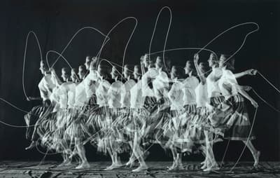 Black-and-white stroboscopic photograph illustrating motion through sequential exposures. A woman wearing a light blouse and patterned skirt moves from left to right while jumping rope. The repeated exposures reveal a continuous wave of her figure alongside dynamic arcs created by the moving rope against a dark background.