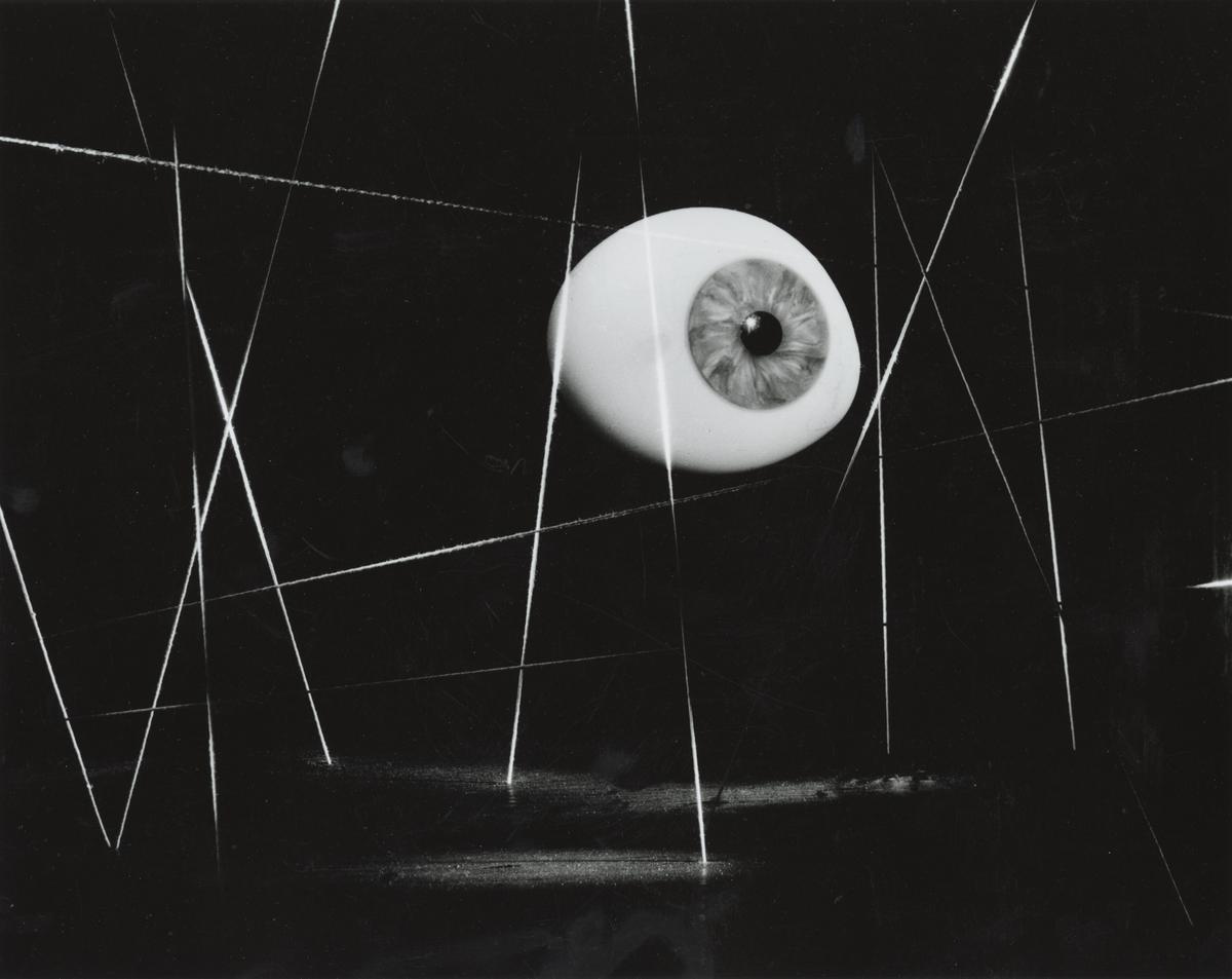 Black-and-white surrealist photograph featuring a single, detailed human eye suspended near the center of a dark, geometric composition. Thin, bright white lines crisscross vertically and horizontally across the black space, anchoring and framing the floating eyeball.