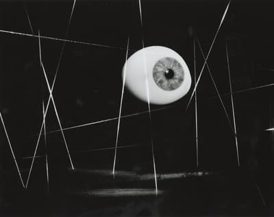 Black-and-white surrealist photograph featuring a single, detailed human eye suspended near the center of a dark, geometric composition. Thin, bright white lines crisscross vertically and horizontally across the black space, anchoring and framing the floating eyeball.