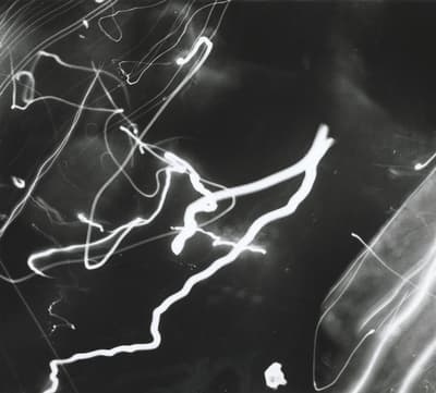 Black-and-white experimental photograph capturing light motion against a dark field. Bright, energetic trails of light loop, zigzag, and curve across the composition, ranging from bold, thick white brush-like strokes to faint, delicate wisps of light.