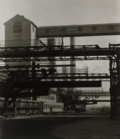 Black-and-white architectural and industrial photograph showing a close view of heavy steel framework, piping, and overhead conveyers. In the background, factory buildings with tall smokestacks rise against a pale sky, emphasizing strong vertical and horizontal geometric lines.