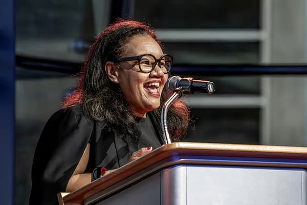 A medium close-up photograph of Erica Wall, director of the Lunder Institute for American Art, speaking behind a wooden podium with a microphone. She is smiling broadly and looking slightly to the right, illuminated by warm stage lighting against a softly blurred background.