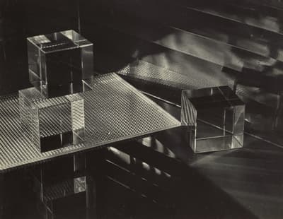 Black-and-white still life photograph depicting transparent geometric cubes resting on grid-patterned and reflective glass planes. Light passes through the objects, creating sharp reflections, shadows, and linear highlights against a dark, dramatic background. Signed and dated in the lower right margin.