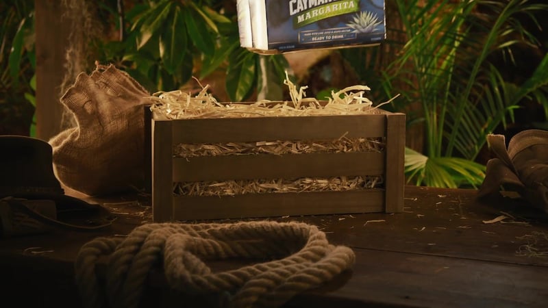 Hands lifting a Cayman Jack bottle from a straw-packed wooden crate in low tropical light