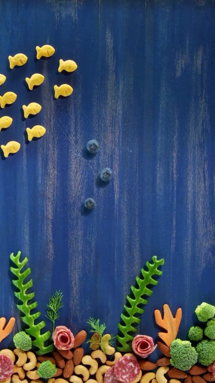 Goldfish crackers arranged as an underwater scene on painted blue board, nuts and green sprigs forming a reef along the bottom