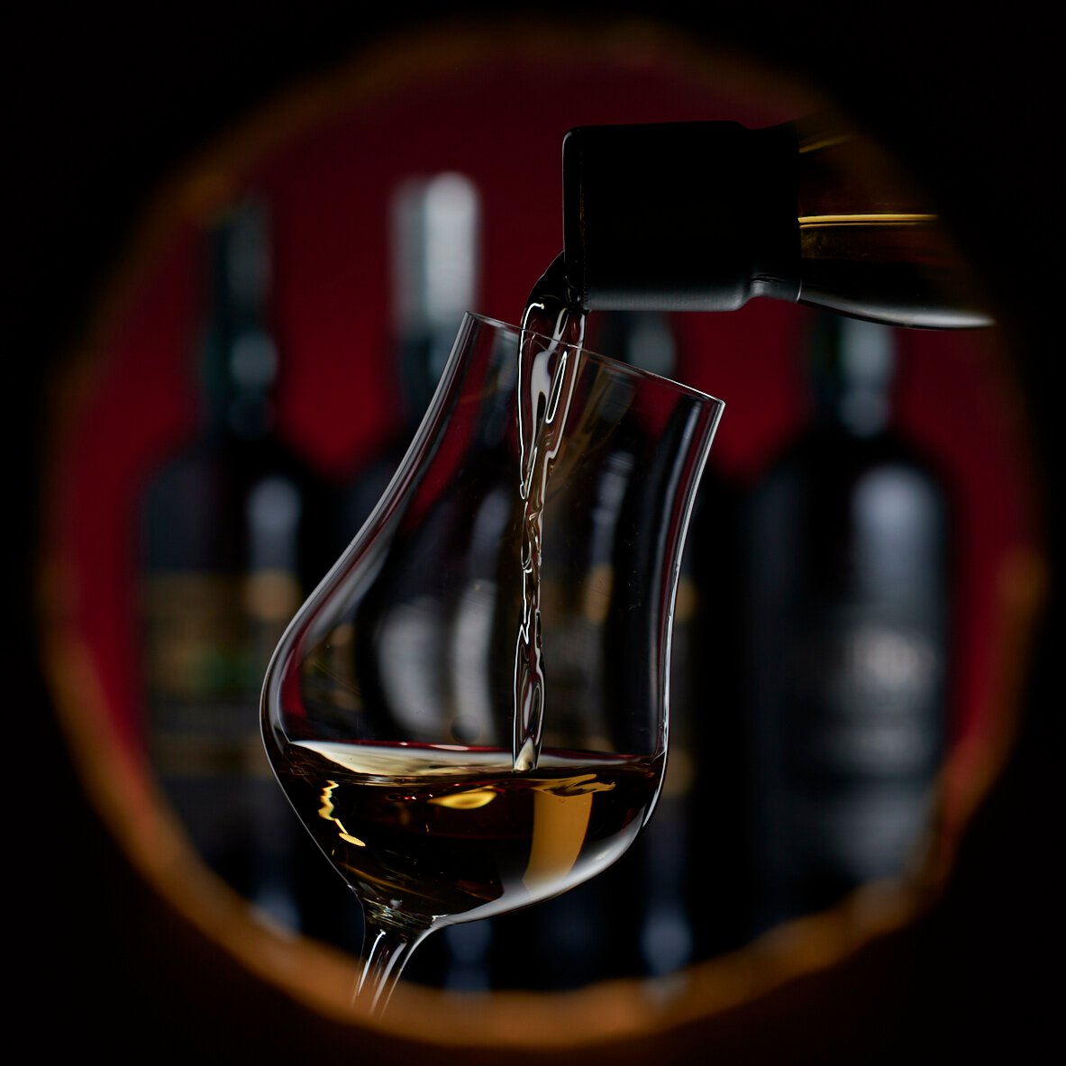 Whisky pours into a nosing glass seen through a round porthole, bottles blurred behind